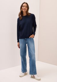 Sweatshirt bleu marine avec des rayures blanches sur les côtés, associé à un jean large bleu clair et des baskets blanches avec des accents rouges.