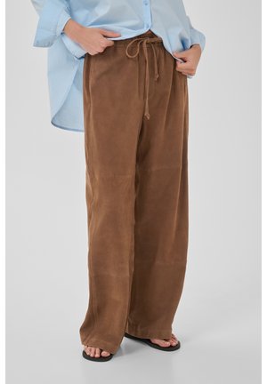 Person trägt lockere braune Kordelzug-Hose, hellblaues Hemd und schwarze Flip-Flops, steht vor schlichtem hellgrauem Hintergrund.