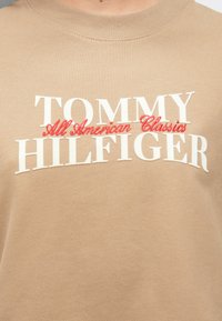 Φυστικί φούτερ με λευκή και κόκκινη κεντητή επιγραφή: "TOMMY HILFIGER All American Classics" στο μπροστινό μέρος. Λείος υφή.