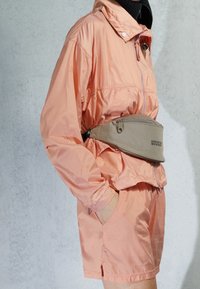 Světle broskvová lehká bunda s vysokým límcem a zipem, doplněná o odpovídající kraťasy. Na pase je obnošená béžová ledvinka.