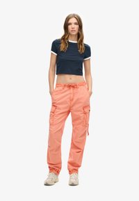 Jonge vrouw staat met het gezicht naar voren in een marineblauw cropped t-shirt, koraalkleurige cargobroek en beige sneakers tegen een witte achtergrond.