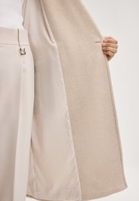 Lichtbeige jas met een zachte, gestructureerde buitenkant en een gladde satijnen voering. Bevat een subtiele naaddetail en een metalen striksluiting op de broek.
