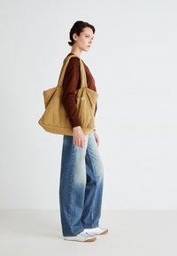 Borsa tote marrone trapuntata con manici larghi e superficie testurizzata, abbinata a jeans in denim azzurro chiaro e sneaker argento.