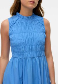 Modrá šaty s nařaseným vysokým límcem, bez rukávů a vlající sukní. Hladká textura s vodorovnými sklady.