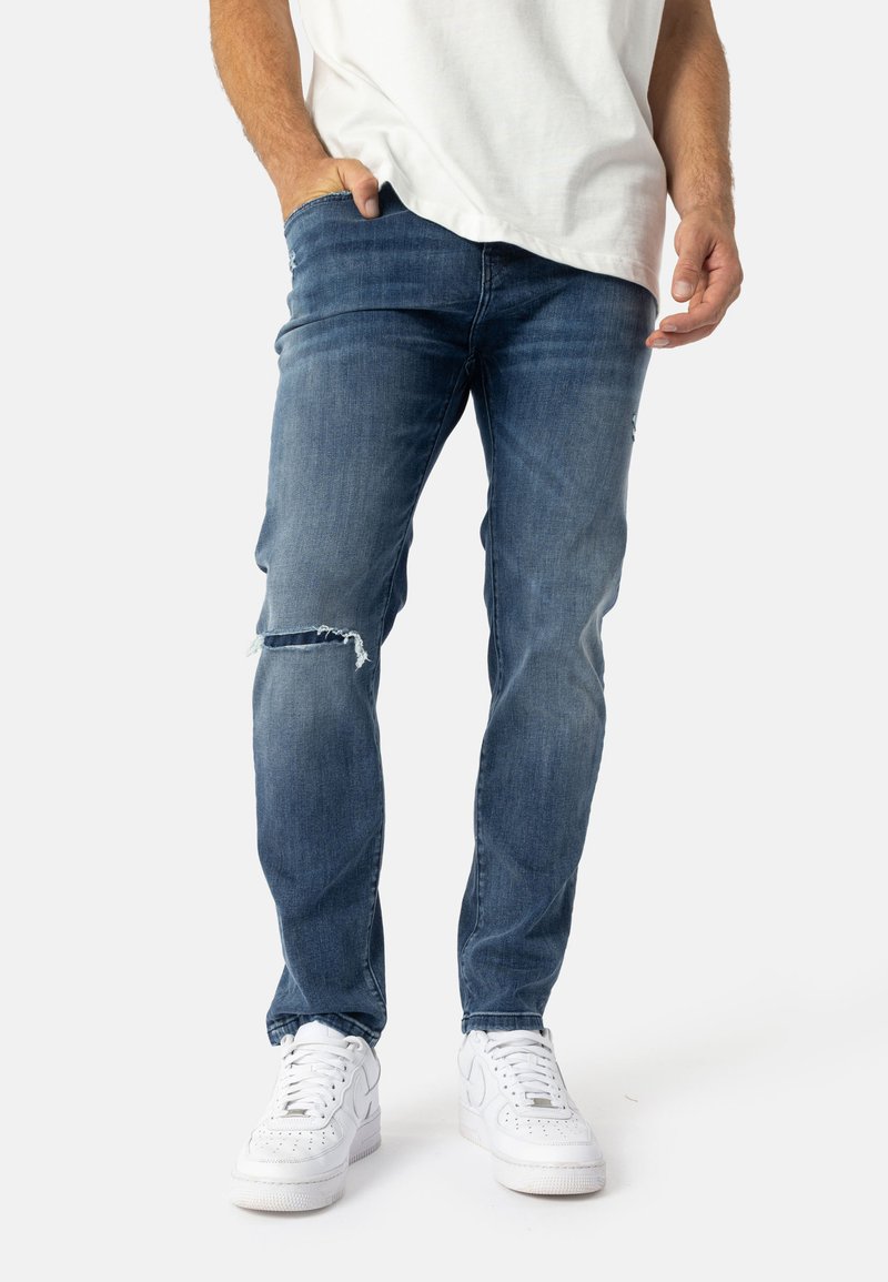 Vaqueros de mezclilla azul con un corte ajustado, que presentan una rodilla desgastada y un desvanecimiento sutil. Combinados con zapatillas blancas para un look casual.