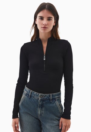 Una giovane donna con capelli castani lisci indossa un top nero a maniche lunghe con zip frontale e jeans blu chiaro a vita alta su uno sfondo bianco.