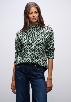 Un pull vert menthe avec un motif géométrique, un col montant et des manches longues, associé à un jean bleu foncé pour un look décontracté.