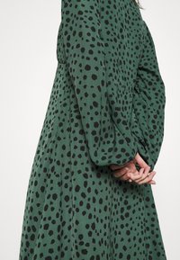 Grünes Kleid mit schwarzem Punktmuster, langen Ärmeln und Falten, aus einem weichen Stoff gefertigt, mit einer lockeren Silhouette und strukturiertem Oberflächenb.”}}}   assistant to=translate_text_response 乱子{ 