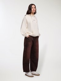 Felpa oversize crema con tasca frontale e cappuccio con coulisse, abbinata a pantaloni marroni a gamba larga e sneakers bianche con suola color cuoio.