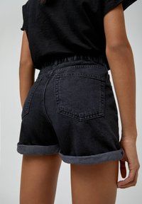 Shorts in denim nero con orli arrotolati, caratterizzati da una vita alta e tasche posteriori, indossati da un modello su uno sfondo neutro.