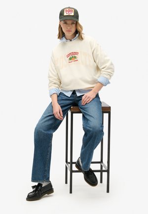 Sudadera crema con el texto "Yosemite" y "Valley Council", superpuesta a una camisa azul clara. Se lleva con jeans azules y zapatos negros. Gorra verde oscuro.
