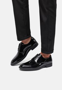Zapatos de vestir de charol negro con punta redonda, diseño con cordones y un acabado suave y brillante, combinados con pantalones oscuros.