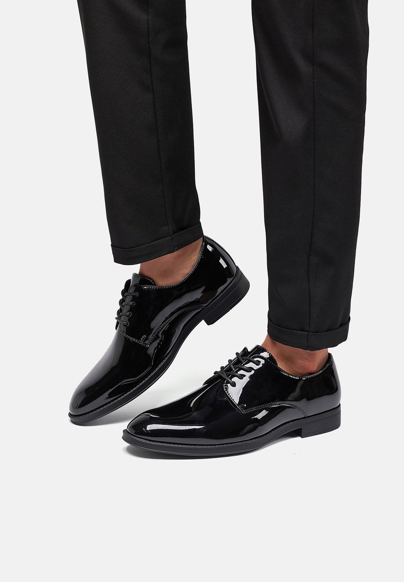Zapatos de vestir de charol negro con punta redonda, diseño con cordones y un acabado suave y brillante, combinados con pantalones oscuros.