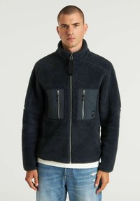 Giacca di shearling blu navy con colletto alto, zip frontale e due tasche sul petto con zip. Superficie tessurizzata con dettagli in tessuto liscio.