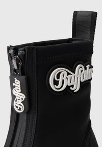 Černé gumové boty na zip, se zdobeným bílým logem a texturovaným švem na horním okraji.