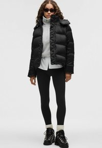 Chaqueta de plumas negra con patrón acolchado, cuello alto y cierre con cremallera. Llevada sobre un suéter gris claro y leggings negros, combinada con botas negras.