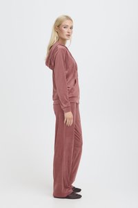 Set di abbigliamento loungewear in velluto color rosa polveroso, composto da una felpa con zip e tasche e pantaloni a gamba larga. Texture morbida, vestibilità comoda, dettagli minimi.