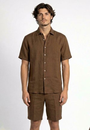 Giovane uomo in piedi, indossa una camicia marrone a maniche corte con bottoni e pantaloncini marroni abbinati su sfondo bianco semplice.