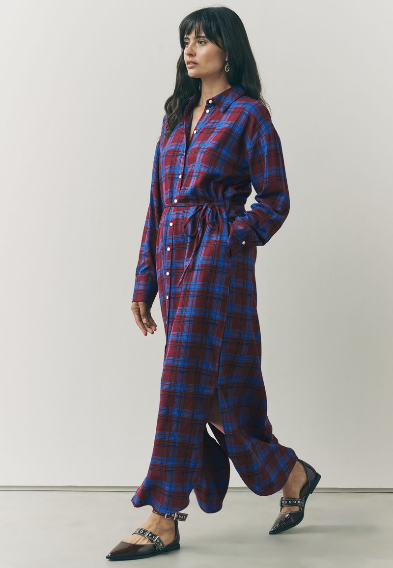 Femme marchant, vêtue d'une longue robe chemise à carreaux rouges et bleus avec une ceinture à la taille, portant des ballerines noires pointues avec des brides autour de la cheville.