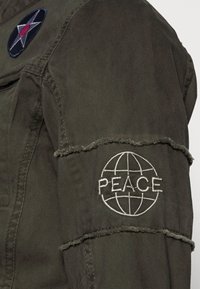 Mouw van een olijfgroene jas met rafelige zoom, voorzien van een ronde geborduurde patch met een sterontwerp en het woord "PEACE" in wit.