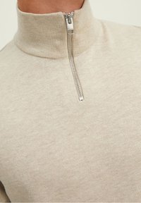 Pull en maille beige avec un col montant côtelé, doté d'une fermeture éclair quart de tour et d'une texture lisse. Les accents comprennent une fermeture éclair en argent.