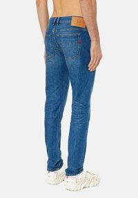 Jeans de denim azul ajustados con un acabado texturizado, costuras rojas en contraste y un parche de cuero marrón en la cinturilla.