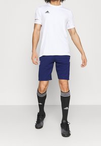 Maglietta atletica bianca a maniche corte con logo nero, abbinata a pantaloni corti blu navy, calze nere fino al ginocchio con strisce bianche e scarpe da calcio nere con tacchetti.