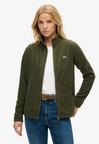 Olive green fleece jacket with a zip front, high collar, two side pockets, and a small brand logo on the chest. Worn over a white shirt.