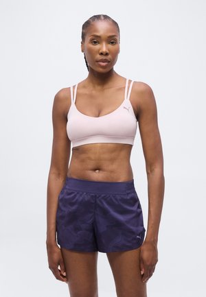Femme aux cheveux tressés portant un soutien-gorge de sport rose clair et un short de sport violet foncé, debout devant un fond blanc uni.