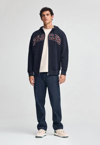 Felpa con zip blu navy con testo "POLO CLUB" in bordeaux, indossata sopra una t-shirt color crema. Abbinata a jeans di denim scuri e sneakers beige.