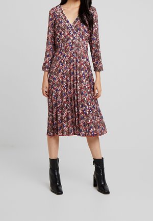 Femme portant une robe portefeuille à fleurs avec des manches trois-quarts et des bottines à talons noirs, debout sur un fond uni.