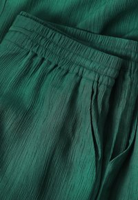 Pantalon vert en tissu texturé avec une ceinture élastique et une poche latérale visible en gros plan.
