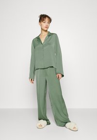 Grön satinpyjamas-set som består av en långärmad skjortblus med knappar och vida byxor. Sätts ihop med vita fluffiga tofflor.