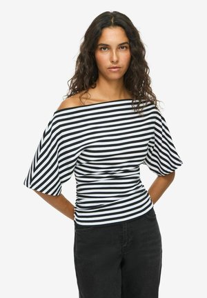 Femme aux longs cheveux bouclés portant un top rayé noir et blanc, épaules dénudées, et un pantalon noir, debout les mains derrière le dos.