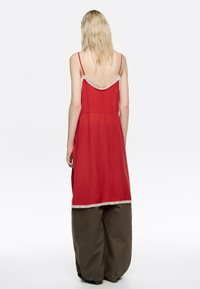 Blondynka w czerwonej sukience bez rękawów z koronkowym wykończeniem, nosząca luźne ciemnobrązowe spodnie, stoi tyłem na białym tle.