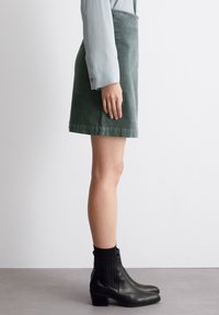 Falda mini de pana verde con corte recto. Se lleva con una blusa de manga larga azul claro y botas de tobillo negras con detalles acanalados.