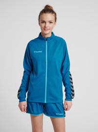 Blauwe sportjas met ritssluiting en een gestructureerde afwerking; voorzien van zwart-witte chevronaccenten op de mouwen. Gecombineerd met bijpassende blauwe shorts.