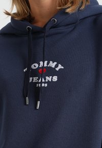Navy blue hoodie with a front graphic reading "TOMMY ♥ JEANS" in white and red. Features drawstring hood and ribbed texture.