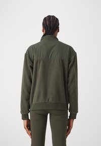 Giacca in pile verde scuro con schiena strutturata, polsini e orlo a costine. Pannello di tessuto liscio nella parte superiore, con design a colletto alto.