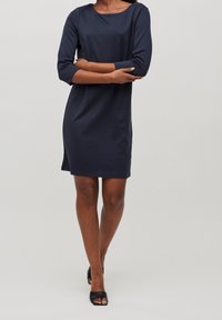 Robe bleu marine, longueur genoux, avec un décolleté bateau et des manches trois-quarts. Tissu lisse avec une légère brillance, sans motifs ni accessoires visibles.