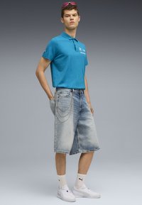 Turkizna polo majica, svetloplave traper šortse sa lančanim detaljem, bele patike i bele čarape sa logotipom. Model pozira protiv gradijentne pozadine.