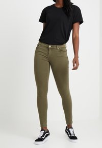 Mujer vestida con una camiseta negra de manga corta, jeans skinny verde oliva y zapatillas blanco y negro, de pie frente a un fondo liso.
