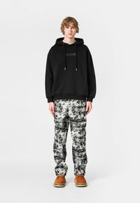 Sudadera con capucha negra y bolsillo frontal, combinada con pantalones blancos y negros con patrón. El modelo lleva calzado marrón. Fondo sencillo.