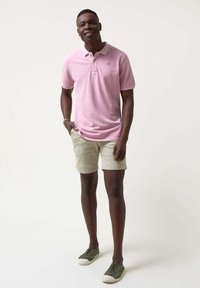Camisa polo rosa de algodón, combinada con shorts beige y zapatillas de lona verdes. Diseño sencillo con un pequeño logo en el pecho.