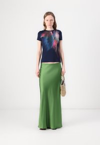 Tengerés t-shirt színes pillangó mintával; zöld selyem maxi szoknya; modell kézben tart egy fonott táskát; sima textúrák és formás sziluettek.