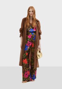 Bruine, lange trenchcoat over een bloeiende jumpsuit met luipaardprint. De jumpsuit heeft krachtige roze, blauwe en rode bloemen. Gouden handtas.