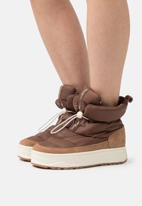 Botas acolchadas marrones hasta el tobillo hechas de tela acolchada con detalles en ante, que cuentan con una suela blanca gruesa y cordones ajustables para un ajuste seguro.