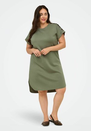 Vrouw draagt een olijfgroene korte mouwen jurk met luipaardprint schouderstrepen en luipaardprint slingback platte schoenen, staand op een witte achtergrond.