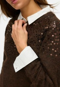 Pull en laine texturée marron avec des accents de sequins sur les épaules, superposé sur une chemise blanche à col. Matière douce et tricotée.