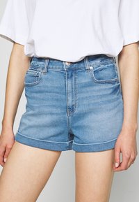 Žena v světle modrých denimových šortkách s ohrnutým lemem a volném bílém tričku, stojící před neutrálním pozadím.